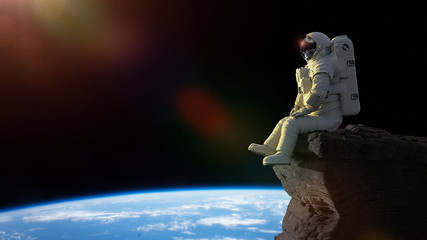 astronaut sitting on a cliff on the Moon in front of planet Earth © dottedyeti