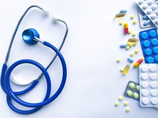 Blue stethoscope, many colorful pills, white, yellow and blue pills in blister packs against a white textured rough paper sheet with place for text