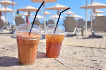 Two refreshing cocktails in plastic cups with black tubes on a wooden table with blurred paid umbrellas on the background. Coffee and ice tea on the beach. Beach holidays with cocktails. 