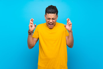 Young man with yellow shirt over isolated blue background with fingers crossing