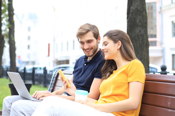 Attrative young couple using laptop computer while sitting on a bench outdoors, holding mobile phone.