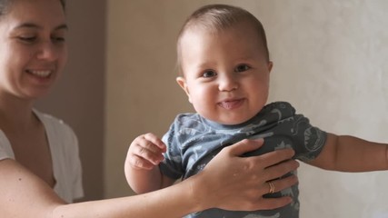 Cute baby doing the first steps and falls in slow motion at home near his mother. Smiles and feels happy