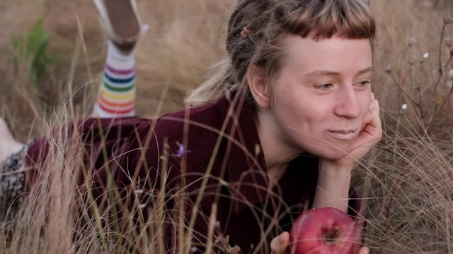 Close look of a girl laying on the wild grass and eating red apple, enjoying summer morning . Summer lifestyle portrait of trendy hipster girl with dreads.