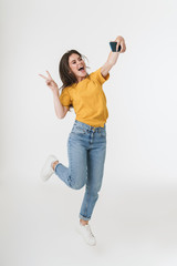 Optimistic young emotional woman jumping isolated over white wall background take a selfie by mobile phone.
