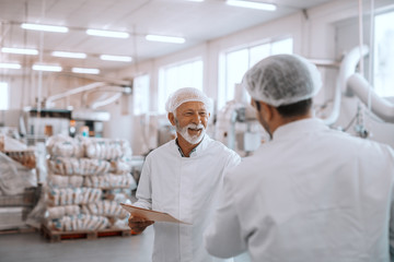 Two supervisors discussing about quality of food in food plant. Older one holding folder with data. Both are dressed in white sterile uniforms and having hairnet.