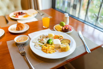 Fresh breakfast table next to window with bread, pastry, egg, fruit, juice, coffee cup