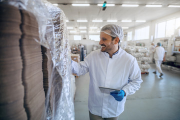 Caucasian smiling employee in white sterile uniform using tablet in food factory.