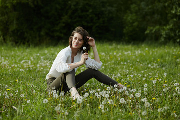 Spring portrait of a girl sitting in a field on the grass among dandelion flowers. Cheerful girl enjoys Sunny spring weather. Natural beauty of a woman, natural cosmetics