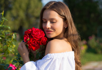 Young beautiful woman with bare shoulders in white in the garden with a red roses.