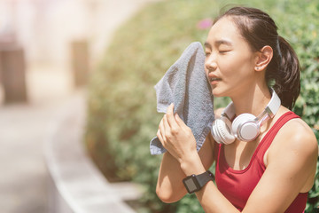 Portrait of tired asian woman sitting on a bench in city park. She is wiping her face using towel after workout session. Horizontal shot