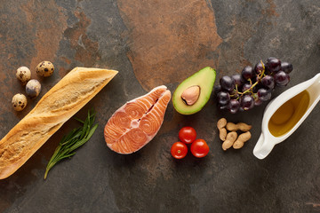 Top view of raw fish near vegetables, fruits, eggs, peanuts, baguette, herb twigs and olive oil on marble surface with copy space