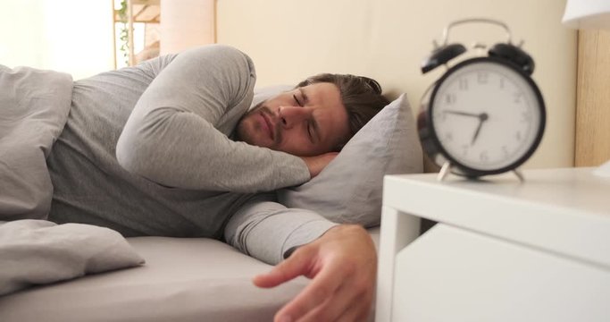 Sleepy man turning alarm clock off and going back to sleep in the morning