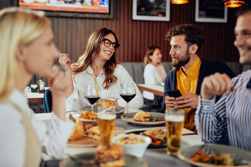 Multiethnic friends sitting at restaurant, drinking alcohol, chatting and having burgers for dinner.
