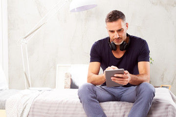 Caucasian man in forties sitting on bed in bedroom and using tablet.