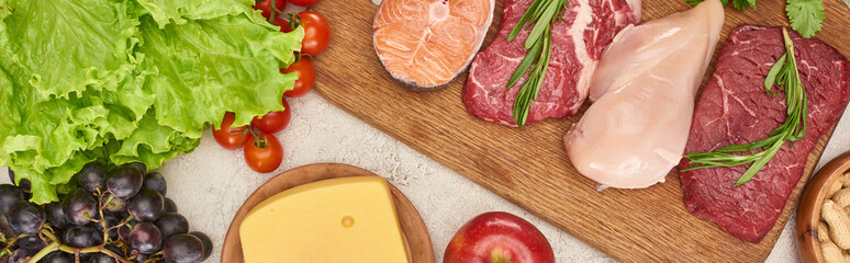Panoramic shot of wooden cutting board with raw fish, meat and poultry near tomatoes, lettuce, cheese and apple on marble surface