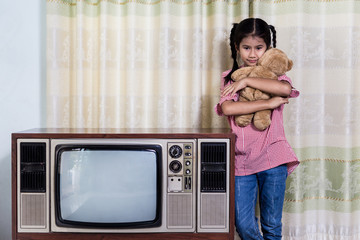 Fototapeta na wymiar Asian little girl is hugging a brown bear and standing with an old TV