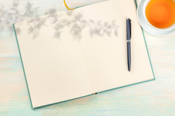 An open journal with an organic shadow, a pen and a cup of tea, shot from the top, on a light...