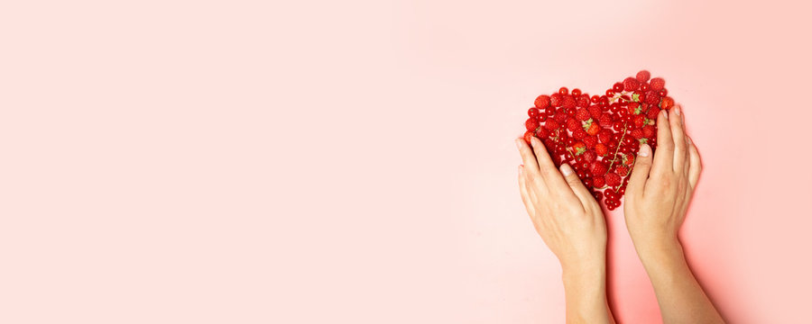 Female hands and red berries in the shape of a heart on a pink background.