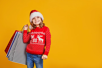 Adorable child in red Santa hat and sweater with deer carrying colorful shopping bags with Christmas gifts on yellow background
