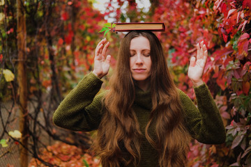 Girl with long hair holding a book on head among colorful ivy in autumn.