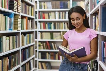Smiling black girl reading book between bookshelves
