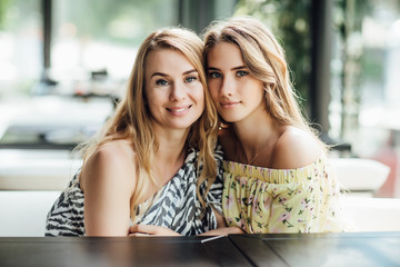 A young beautiful blonde girl and her mom rest on a summer terrace cafe, communicate. They are happy and funny.