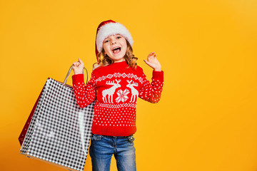 Adorable child in red Santa hat and sweater with deer carrying colorful shopping bags with Christmas gifts on yellow background