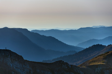 A small man (on the left) admires a series of valleys covered by forest in the evening light giving the scene a sense of scale