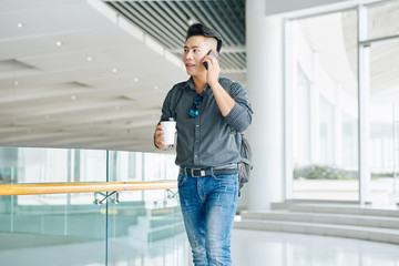 Young Asian man walking in office building and talking on hone with coworker or clienthe is going to meet with