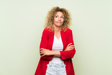 Young blonde woman with curly hair over isolated green background making doubts gesture while lifting the shoulders