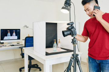 Young Vietnamese photographer calling on phone when setting up digital camera for shooting in his small home studio © DragonImages