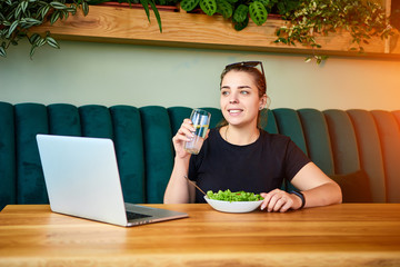 Young happy woman drinks water in the beautiful interior with green flowers on the background and fresh ingredients on the table. Healthy food concept