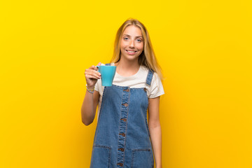 Blonde young woman over isolated yellow background holding hot cup of coffee