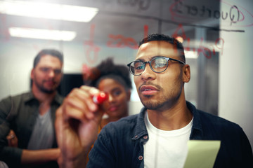 Business team looking at young businessman writing ideas and plans down on glass wall sticky notes and ideas