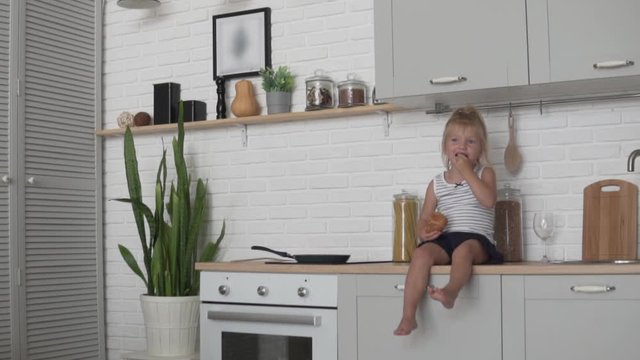 Funny little girl sitting in the kitchen and eagerly eating a loaf of white bread. A child sits in the kitchen with a baguette of bread