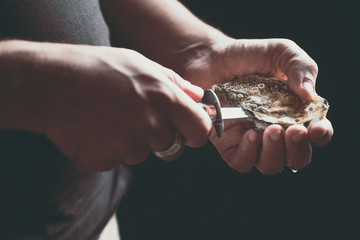 A man with a knife opens a fresh oyster, a drop of water on his hand. Dark rustic background, selective focus. Close-up