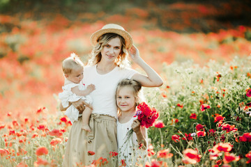 mother holds her hat, baby looks at her sister on the poppy field