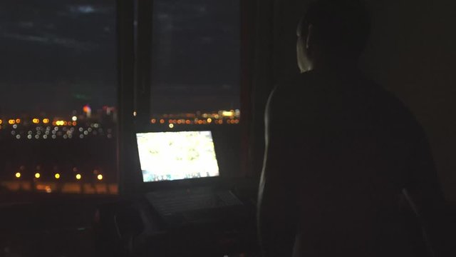 Fat young man movements on the treadmill and works by notebook at night by the panoramic window with bokeh lights and amazing urban city view