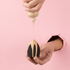 Girl hand holds a beauty blender in foundation, concealer from a hole in a pink background. Makeup artist concept, copy space.