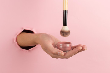 Girl hand holds a brush for makeup and powder from a hole in a pink background. Makeup artist concept, copy space.