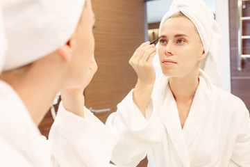 Close-up young well-groomed slender girl adjust eyebrows after taking a bath, stands with a towel and bathrobe in front of large young mirror. Beautiful woman getting ready to go out and doing makeup
