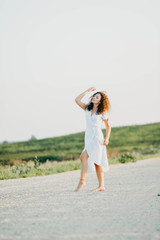 curly young woman dancing romantically in a blue dress on a sandy road at sunset.