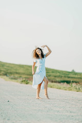 curly young woman dancing romantically in a blue dress on a sandy road at sunset.