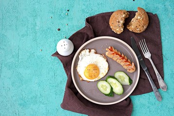 Breakfast, fried egg with sausage and slices of fresh cucumbers on a brown clay plate on a turquoise concrete background. Children's breakfasts. School breakfast concept. Top view, copy space.