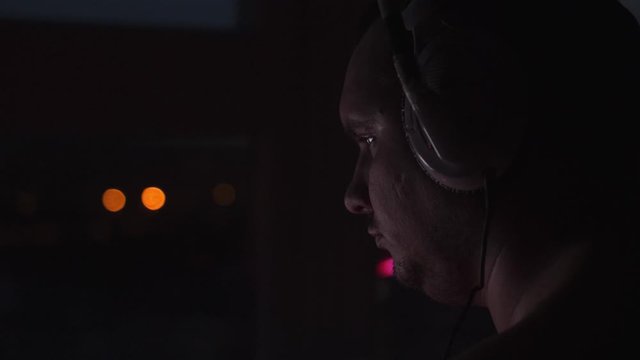 Close up of man in headphones playing video game at home in dark room at night enjoying gaming activity.