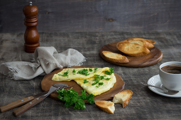 Morning breakfast of a traditional French omelet with toasts and butter, chopped parsley and a white porcelain cup of coffee on a wooden board placed on a dark wooden table, top view