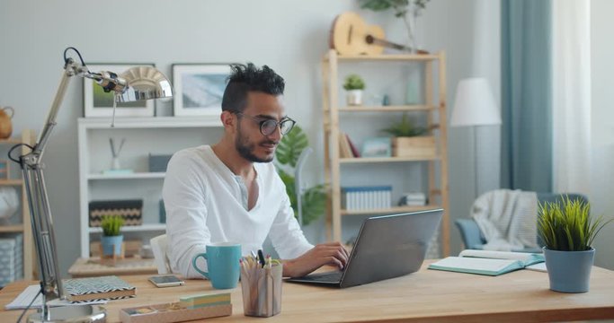 Portrait of handsome young Arab using laptop at home then smiling looking at camera sitting at table alone. People, lifestyle and remote work concept.
