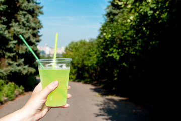 Woman holding plastic green cocktail cup close-up, outdoors in summer in the park