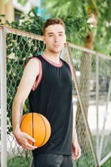 Positive handsome young man in black sports uniform standing at net around outdoor basketball court...