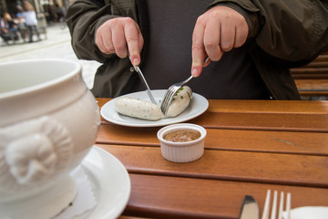 Mann isst Münchener Weißwurst mit Messer und Gabel, süßen Senf und Brezel und löst Wurst richtig aus Pelle draußen in Biergraten